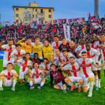 Giocatori del Genoa in festa con sciarpe e bandiere rossoblù davanti alla tifoseria: celebrazione nel derby Pisa–Genoa dopo la vittoria.
