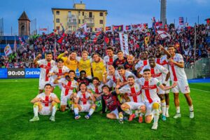 Giocatori del Genoa in festa con sciarpe e bandiere rossoblù davanti alla tifoseria: celebrazione nel derby Pisa–Genoa dopo la vittoria.