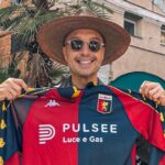 Sorriso davanti a una facciata storica con una maglia Genoa rosso-blu PULSE, sciarpa/cappello di paglia e occhiali da sole