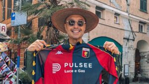 Sorriso davanti a una facciata storica con una maglia Genoa rosso-blu PULSE, sciarpa/cappello di paglia e occhiali da sole