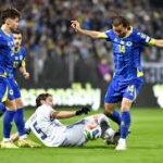 Italia contro Bosnia Erzegovina: giocatori in maglie blu si contengono sul campo verde, pubblico sullo sfondo, tensione e contesa sportiva tra le squadre.