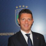 Gianluca Rocchi in abito scuro e cravatta blu davanti a logo FIGC, nel servizio sul designatore Rocchi indagato per frode sportiva e autosospensione a Carrara