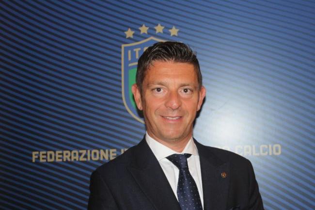 Gianluca Rocchi in abito scuro e cravatta blu davanti a logo FIGC, nel servizio sul designatore Rocchi indagato per frode sportiva e autosospensione a Carrara