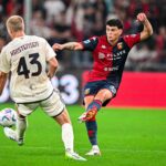 Alan Matturro in maglia rossa e blu con braccio alzato in campo, mentre affronta un avversario: scena di calcio della rubrica sui prestiti Genoa.