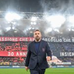 Allenatore del Genoa in abito scuro sul campo: Daniele De Rossi davanti a uno stadio notturno con fumogeni e curva rosso-blu, tra scritte celebrative.