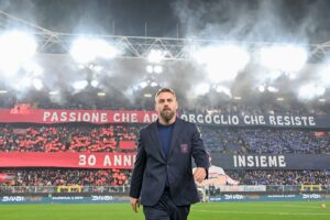 Allenatore del Genoa in abito scuro sul campo: Daniele De Rossi davanti a uno stadio notturno con fumogeni e curva rosso-blu, tra scritte celebrative.